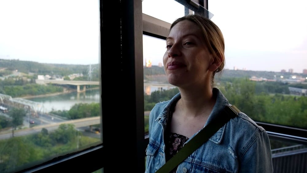 Audrey Bergner stands inside the glass cabin of Edmonton’s 100 Street Funicular, with the North Saskatchewan River valley, bridges and city skyline visible through the windows.