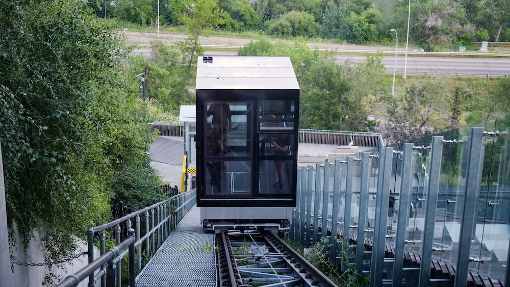 The glass cabin of Edmonton’s 100 Street Funicular descends along the sloped track toward the river valley, with trees, the lower platform and roadway visible below in Edmonton, Alberta, Canada.