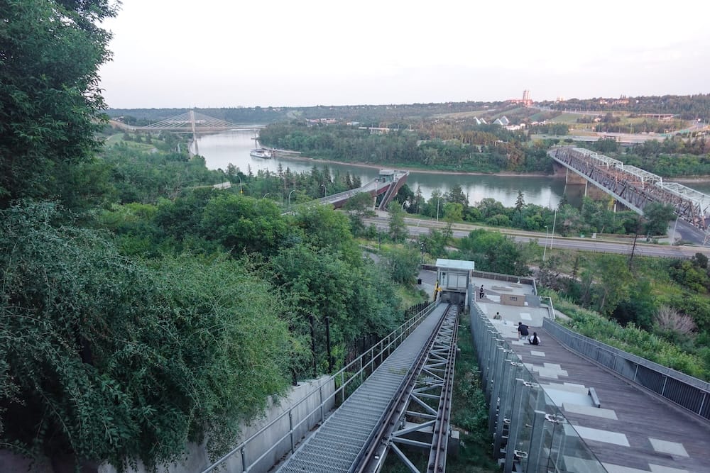 View down the 100 Street Funicular in Edmonton, Alberta, Canada, with the sloping track and promenade leading toward the North Saskatchewan River, multiple bridges and the Muttart Conservatory in the distance.