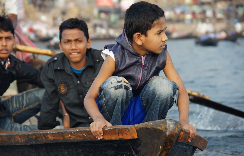 A close-up telephoto shot of a group of young Bangladeshi boys/teenagers on a small river boat plying the waters of the Buriganga.