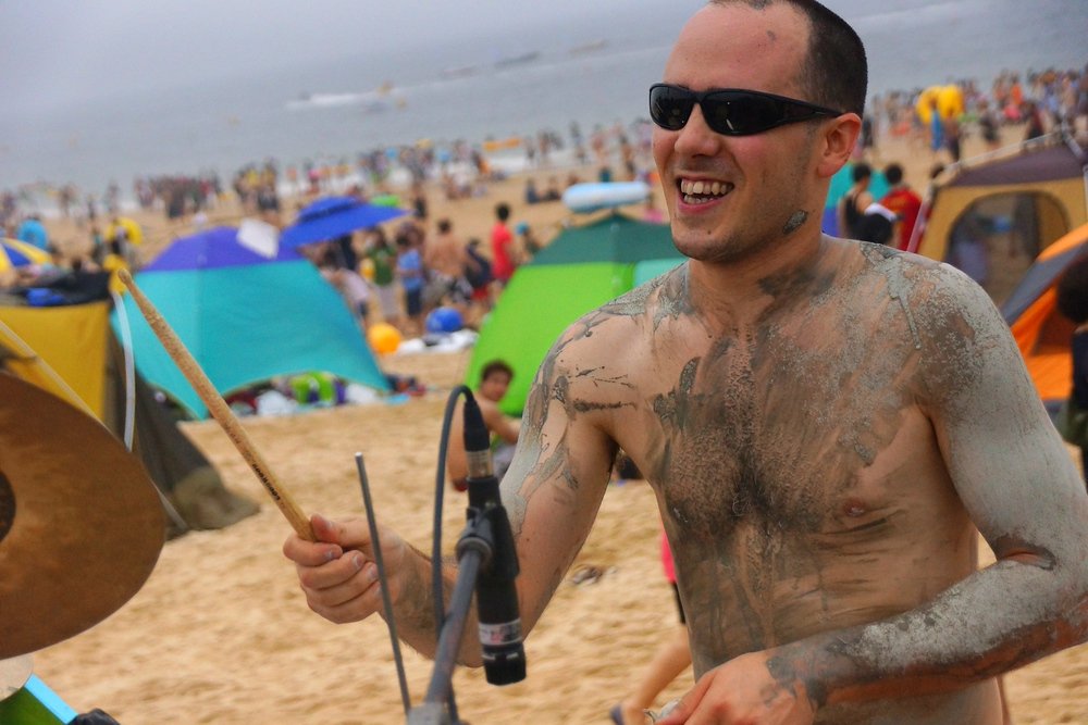 A drummer plastered in mud without a shirt smiles while entertaining thousands with his slick skills during the Boryeong Mud Festival