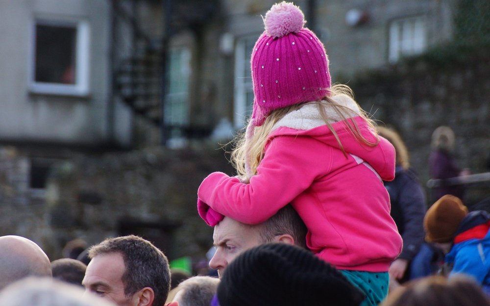 A girl wearing a warm pink fleece and toque sits on top of her Dad's shoulders while watching the Loony Dook in Edinburgh, Scotland.