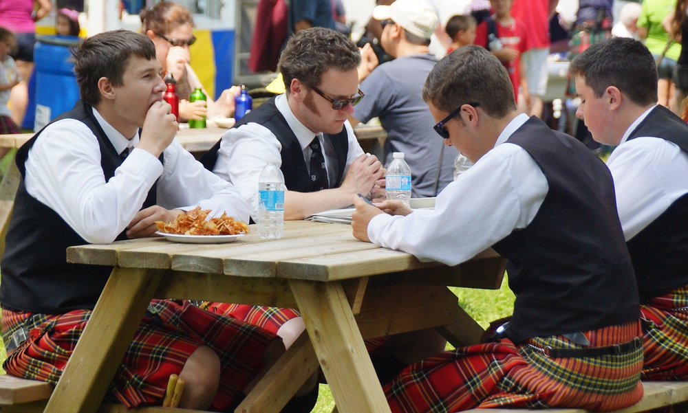 A group of kilted young men and teenagers sit down on a picnic bench to enjoy some snack food at the New Brunswick Highland Games