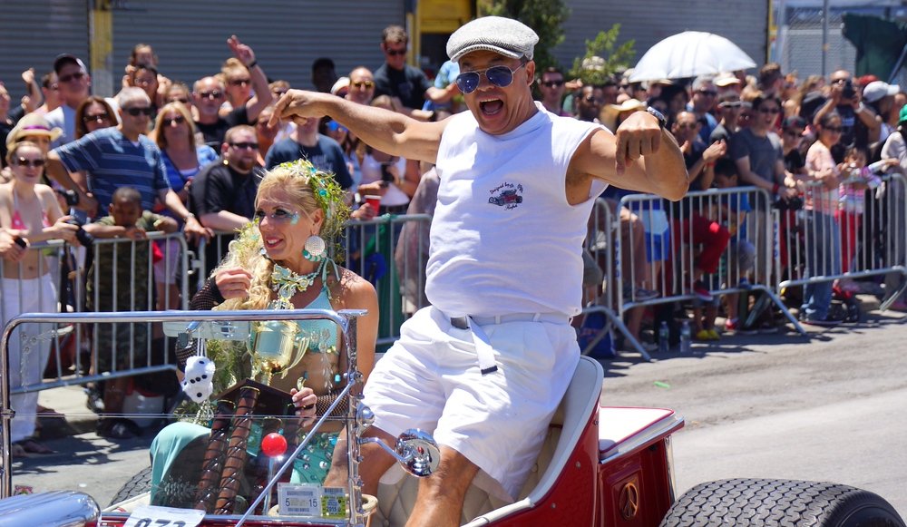 A highly animated Mermaid parader driving a nice racing car down the street in New York City