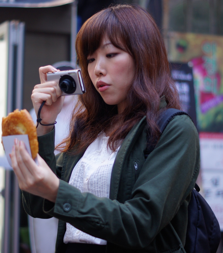 A lady takes a photo of the street food she has just bought by extending her left arm and holding her point and shoot in the other in Insadong, Seoul, Korea