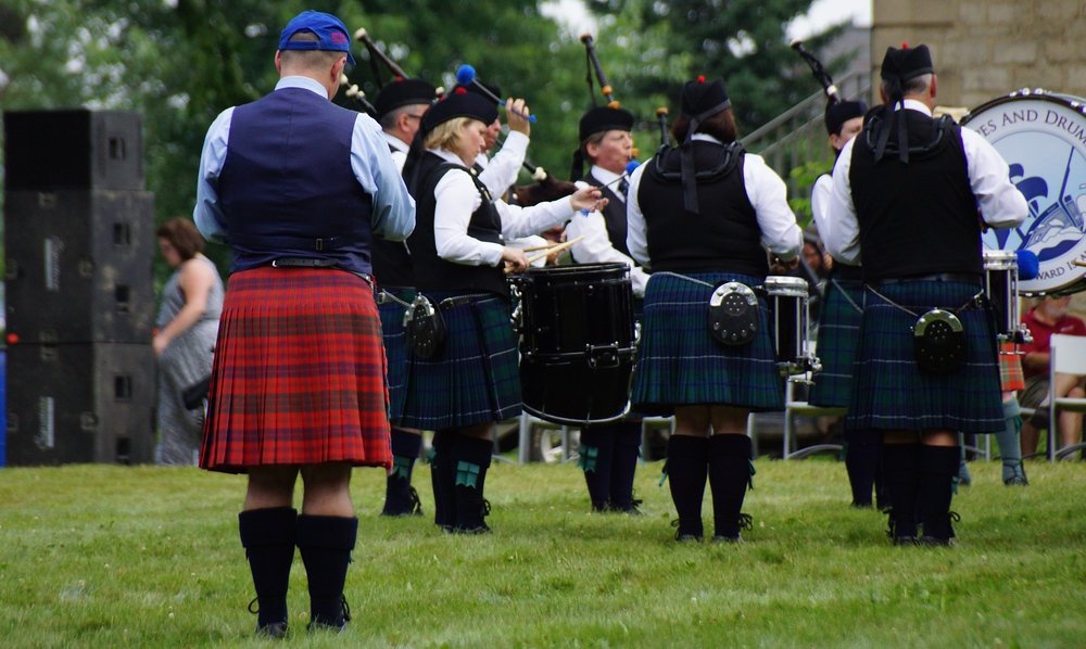 A lone man wearing a red kilt stands behind a band wearing different style kilts at the New Brunswick Highland Games