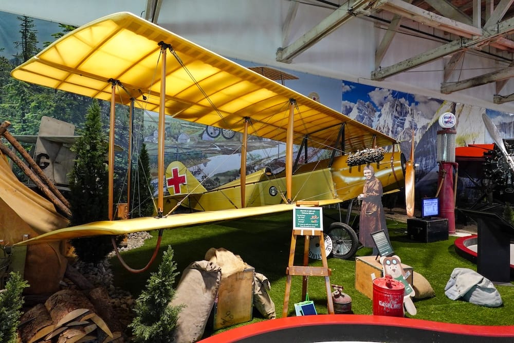 Yellow historic biplane with a red cross marking displayed inside the Alberta Aviation Museum in Edmonton, Alberta, Canada, surrounded by aviation props, painted scenery and hangar beams.