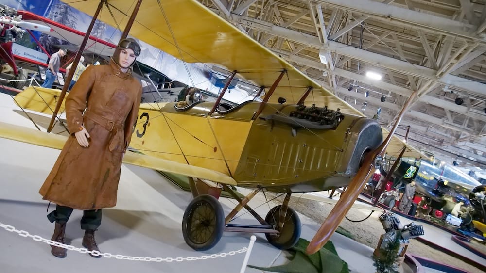 Un biplano amarillo clásico de cabina abierta con hélice de madera y motor expuesto junto a un maniquí vestido con equipo de vuelo de cuero de la época en el Alberta Aviation Museum en Edmonton, Alberta, Canadá.
