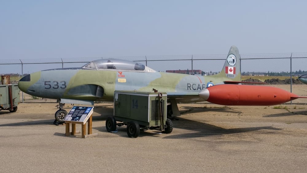 Un jet de entrenamiento clásico de la RCAF pintado de camuflaje con cabina en tándem y marcas de escarapelas canadienses en exhibición al aire libre detrás de un cerco de alambre tejido en el Alberta Aviation Museum en Edmonton, Alberta, Canadá.