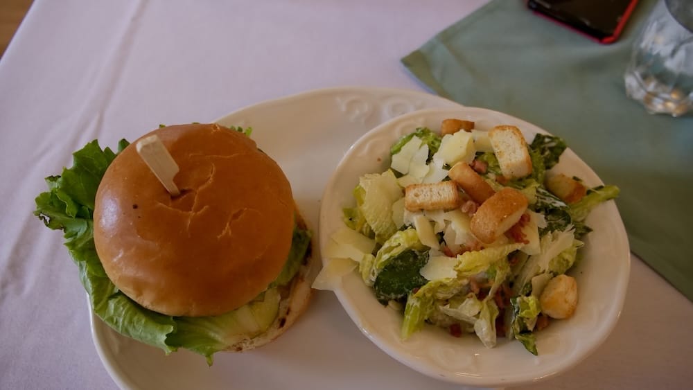 An Alberta beef burger topped with lettuce beside a bowl of Caesar salad with croutons, shaved parmesan, and bacon, served at the Wainwright Hotel at Heritage Park in Calgary, Alberta, Canada.