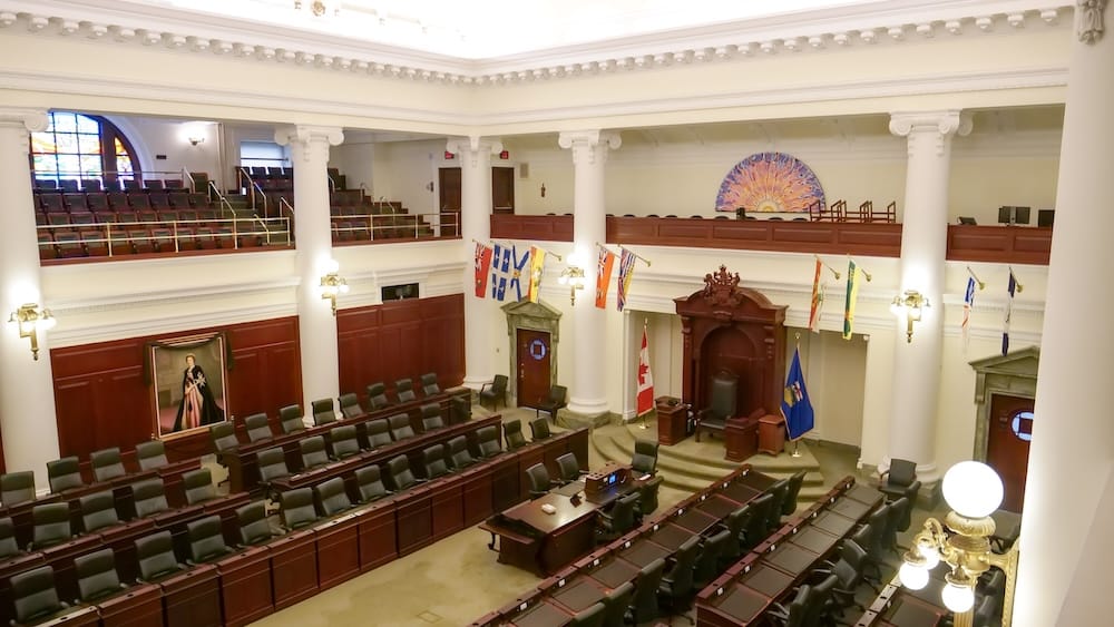 Alberta Legislature Assembly Chamber interior with columns and legislative seating in Edmonton, Alberta, Canada Interior view of the Alberta Legislature Assembly Chamber in Edmonton, Alberta, Canada, showing rows of legislative seating, tall white columns, wood panelling, flags and the Speaker’s chair.