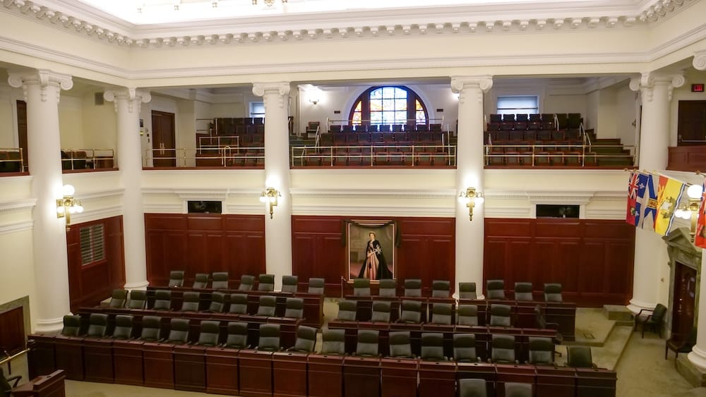 Interior of the Alberta Legislature assembly chamber in Edmonton, Alberta, Canada, with rows of desks, white columns, wood panelling, provincial flags and a portrait beneath the gallery.