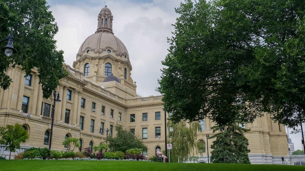 Alberta Legislature Building in Edmonton, Alberta, Canada, with its domed sandstone exterior rising behind landscaped lawns, mature trees, flower beds and a seated visitor on the government grounds.