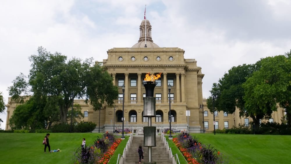 Alberta Legislature Building in Edmonton, Alberta, Canada, framed by green lawns and flower beds, with the domed sandstone façade and a ceremonial flame burning in front.