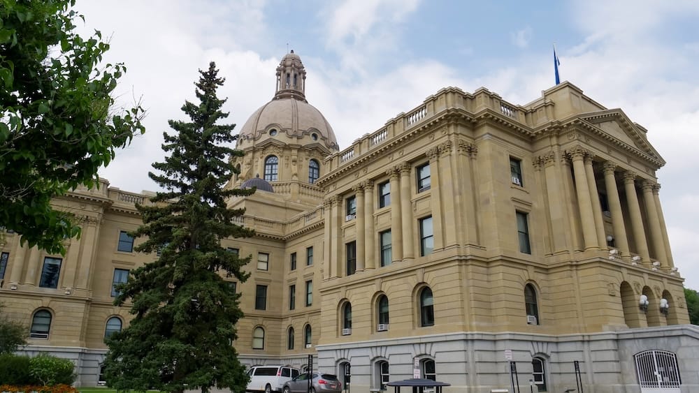 Alberta Legislature Building, Edmonton, Alberta, Canada — the provincial capital's domed sandstone landmark. The Alberta Legislature Building in Edmonton, a Beaux-Arts sandstone landmark with a domed cupola, photographed from the grounds beside a tall evergreen tree under a partly cloudy sky, marking Alberta's provincial capital.