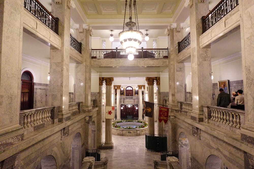 Interior of the Alberta Legislature Building in Edmonton, Alberta, Canada, with marble columns, ornate railings, chandeliers and visitors overlooking the grand central hall.