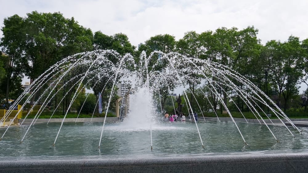 Large arcing fountain in front of the Alberta Legislature grounds in Edmonton, Alberta, Canada, with water jets forming a dome above the pool and leafy trees behind.