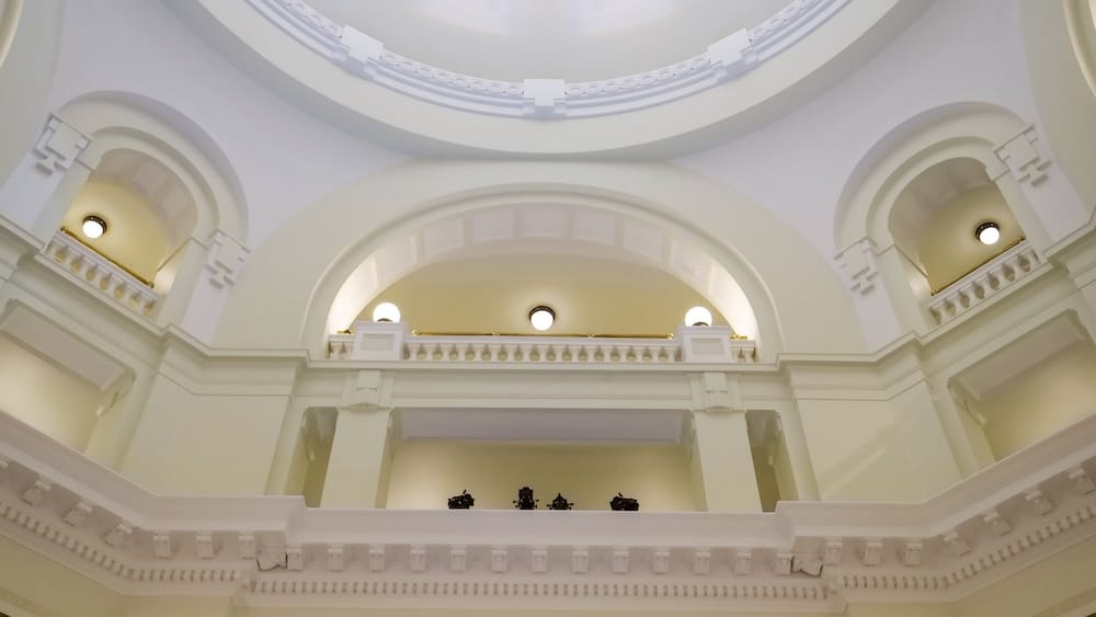 Interior view looking upward inside the Alberta Legislature Building in Edmonton, Alberta, Canada, showing the pale domed ceiling, broad arches, decorative balconies and glowing circular lights.