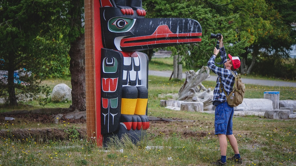 Nomadic Samuel Creatively Capturing Indigenous Totem Pole Photography in Alert Bay, British Columbia Alert Bay, British Columbia — Nomadic Samuel raising his camera to photograph a colorful Indigenous totem pole, capturing original creative travel photography and behind-the-scenes content for Nomadic Samuel destination guides, videos, and licensing use.