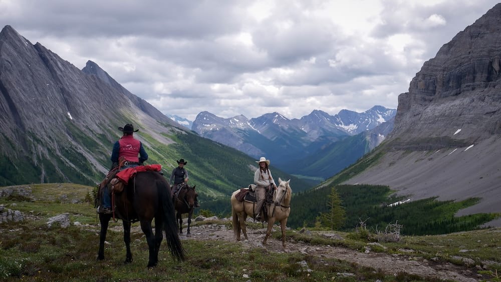 Allenby Pass, Banff National Park, Alberta, Canada - backcountry horseback riders above a mountain valley Three riders on horseback pause at Allenby Pass in Banff National Park, Alberta, Canada, with steep grey mountain walls, a green valley and layered Rocky Mountain peaks under a cloudy sky.