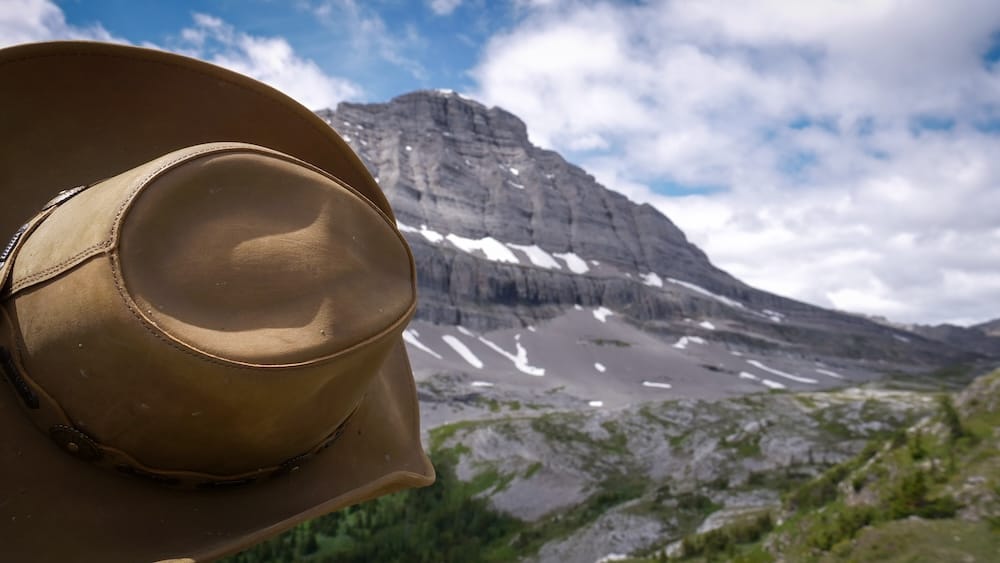 Un sombrero cowboy marrón ocupa el primer plano en Allenby Pass, Parque Nacional Banff, Alberta, Canadá, con picos rocosos en capas, restos de nieve y terreno alpino extendiéndose al fondo.