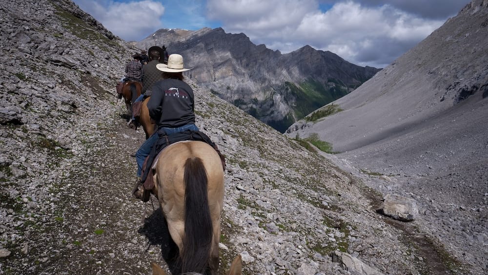 Horseback riders climbing toward Allenby Pass in Banff National Park, Alberta, Canada on a rocky alpine trail Horseback riders climb a narrow rocky trail toward Allenby Pass in Banff National Park, Alberta, Canada, with steep scree slopes and layered gray mountain walls surrounding the alpine route.