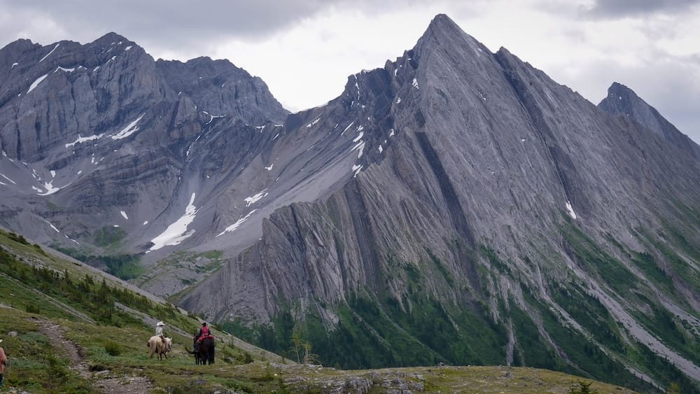 Tiny horseback riders cross the high-alpine terrain at Allenby Pass in Banff National Park, Alberta, Canada, beneath enormous jagged grey mountain peaks with lingering patches of snow.