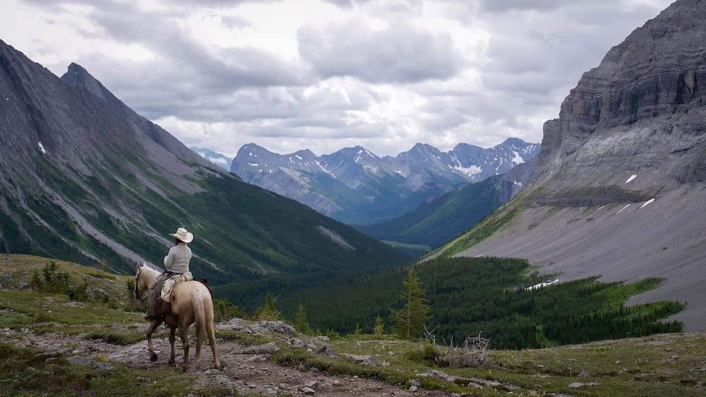 Horseback riding at Allenby Pass in Banff National Park, Alberta, Canada with alpine valley and mountain scenery A horseback rider travels across Allenby Pass in Banff National Park, Alberta, Canada, overlooking a deep forested valley framed by steep rocky mountains and distant snow-streaked peaks.