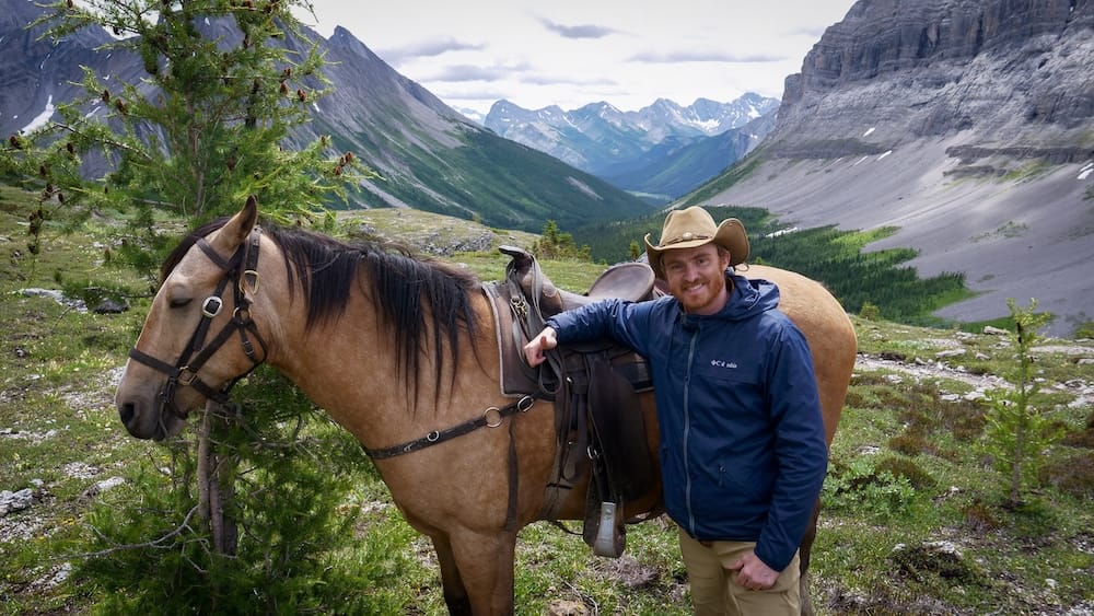 Samuel Jeffery with horse at Allenby Pass, Banff National Park, Alberta, Canada backcountry horseback ride Samuel Jeffery stands beside a saddled horse at Allenby Pass in Banff National Park, Alberta, Canada, with an alpine valley, rocky slopes and layered mountain peaks stretching into the distance behind him.