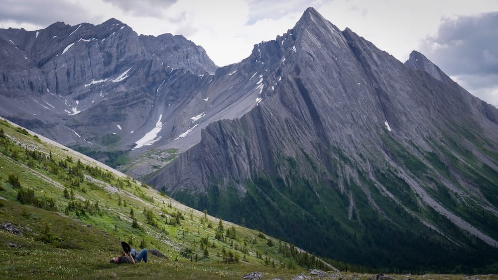 Two tiny people rest on a grassy slope at Allenby Pass in Banff National Park, Alberta, Canada, while jagged grey peaks and steep alpine walls dominate the landscape, emphasizing the immense scale of the backcountry scenery.