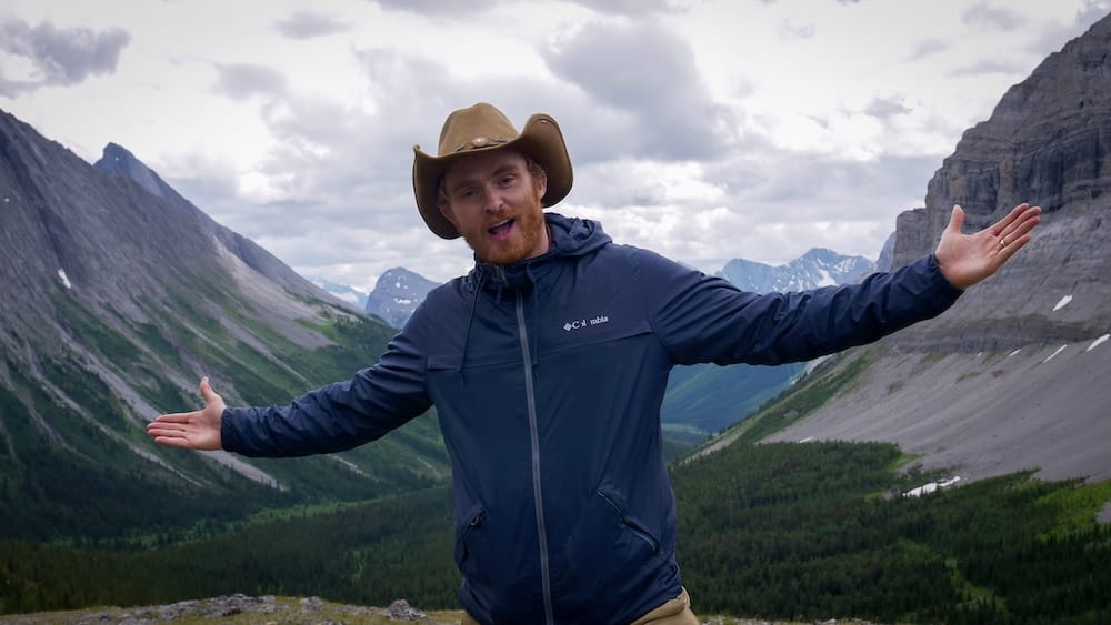 Samuel Jeffery stands at Allenby Pass in Banff National Park, Alberta, Canada, wearing a cowboy hat with his arms outstretched as steep alpine slopes and a deep mountain valley fill the background.