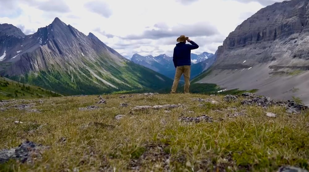 Samuel Jeffery stands on a grassy alpine slope at Allenby Pass in Banff National Park, Alberta, Canada, looking out over a broad backcountry valley framed by steep grey mountain walls and jagged peaks.