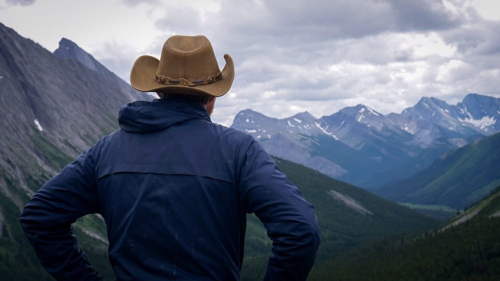 Samuel Jeffery, visto de espaldas con sombrero cowboy y campera azul, contempla el valle montañoso desde Allenby Pass en el Parque Nacional Banff, Alberta, Canadá, con picos en capas y laderas boscosas extendiéndose a la distancia.