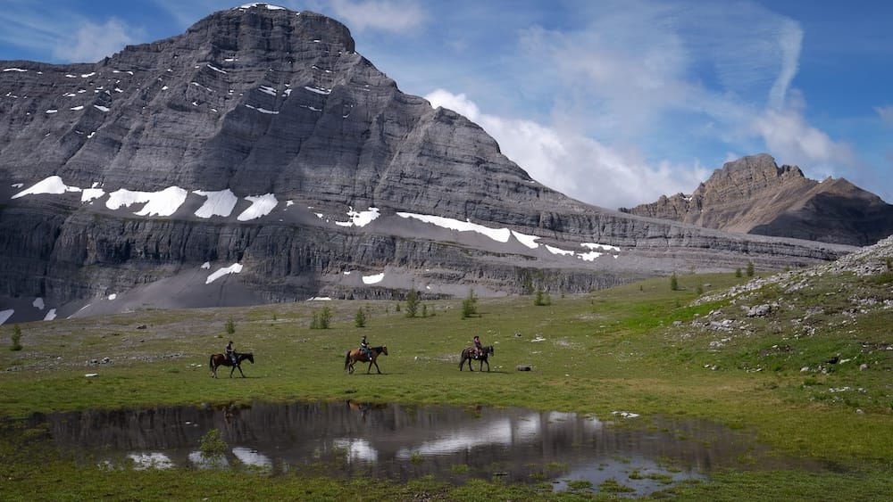 Ruta a caballo por la zona agreste cerca del paso Allenby, Parque Nacional Banff, Alberta, Canadá Jinetes a caballo cruzando una pradera alpina remota cerca de un pequeño lago glacial en la ruta al paso Allenby en el Parque Nacional Banff, Alberta, Canadá, con una montaña veteada de nieve a sus espaldas.