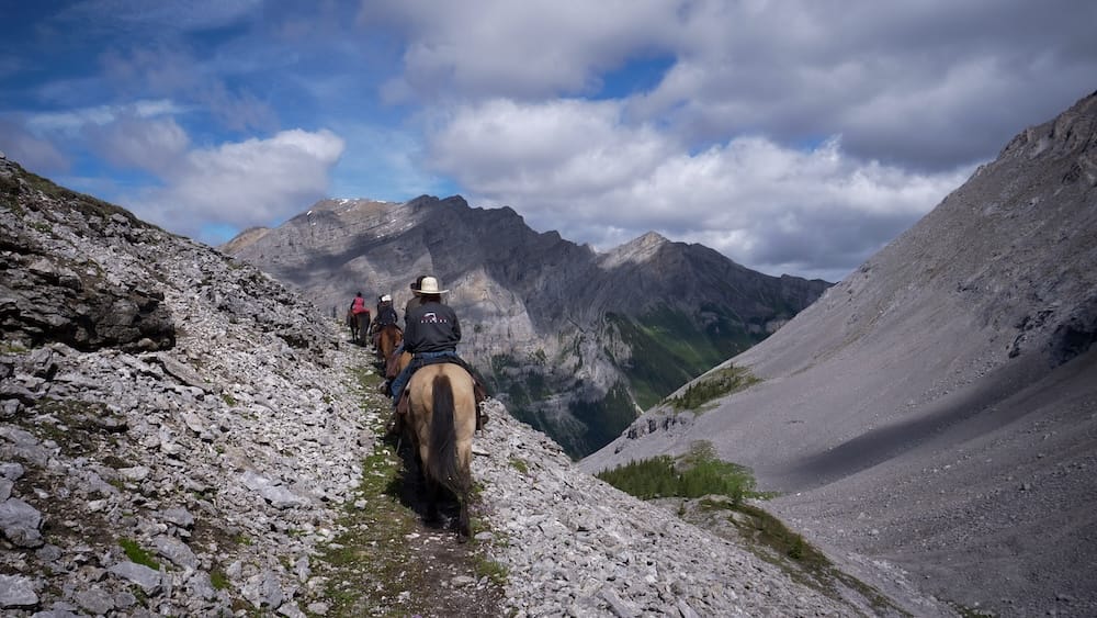 Jinetes avanzan por un sendero angosto y rocoso hacia Allenby Pass en el Parque Nacional Banff, Alberta, Canadá, con laderas de pedregal, paredones montañosos expuestos y un valle profundo abriéndose adelante.
