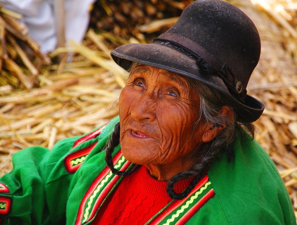 An elderly lady with leathery skin and wrinkles wearing a bowler hat and colourful attire from Lake Titicaca, Peru 