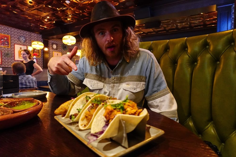 Samuel Jeffery con tacos en el restaurante mexicano Añejo de Banff, Alberta, Canadá Samuel Jeffery está sentado en un box de Añejo en Banff, Alberta, Canadá, señalando un plato de tacos sobre la mesa, con platos de salsa al costado y el tapizado verde capitoné del restaurante detrás.