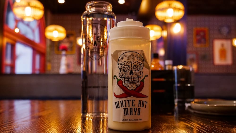 White Hot Mayo condiment at Añejo Restaurant in Banff, Alberta, Canada Bottle of White Hot Mayo on a table inside Añejo in Banff, Alberta, Canada, featuring a sugar-skull and red-chili label with warm pendant lights and restaurant decor in the background.