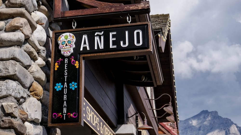Añejo Restaurant exterior sign on Banff Avenue in Banff, Alberta, Canada Exterior Añejo Restaurant sign in Banff, Alberta, Canada, mounted beside a stone facade with a cloudy mountain backdrop visible beyond the building.