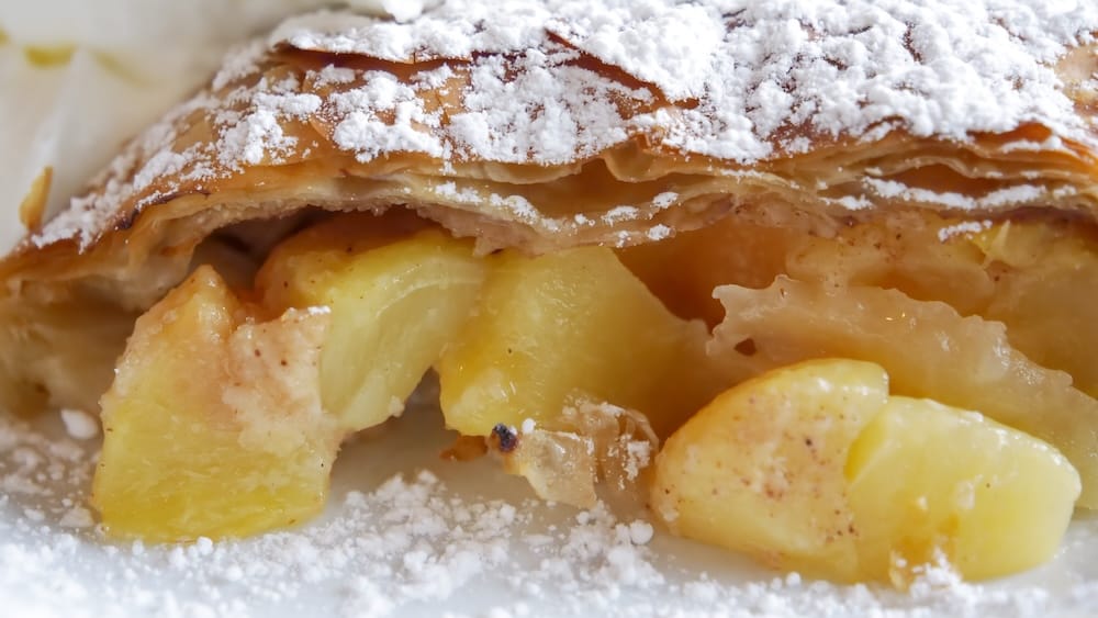 Primer plano del strudel de manzana austríaco en Bistro Praha en Edmonton, mostrando la masa hojaldrada espolvoreada con azúcar impalpable y rodajas de manzana asada suave en su interior.