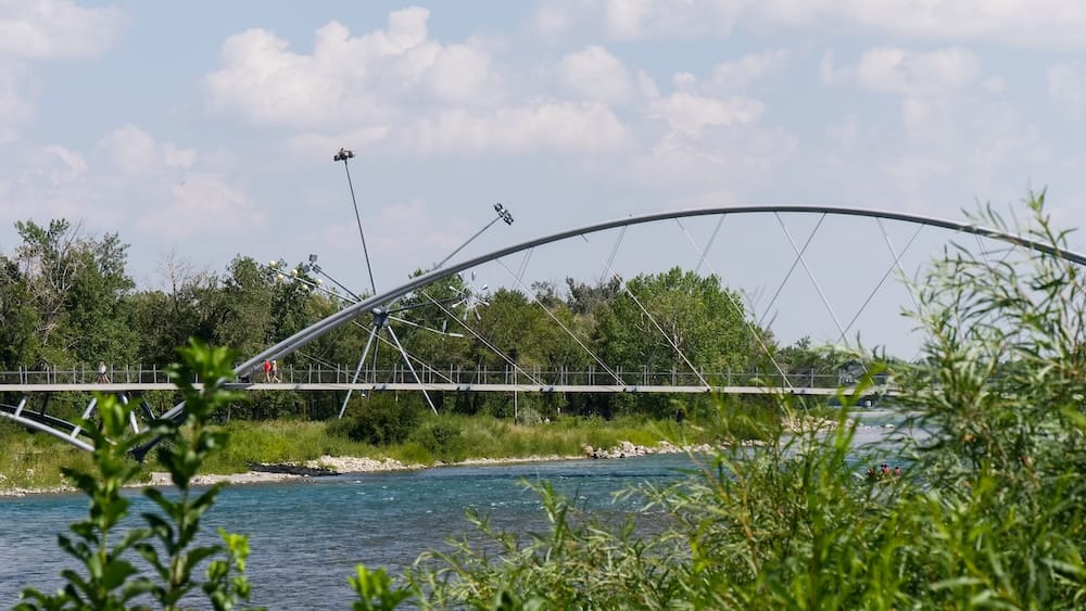 Arched pedestrian bridge along the Bow River, Calgary, Alberta, Canada A silver arched pedestrian bridge spans the Bow River along the RiverWalk pathway in Calgary, Alberta, Canada, with people crossing on foot and rafters visible in the water below.