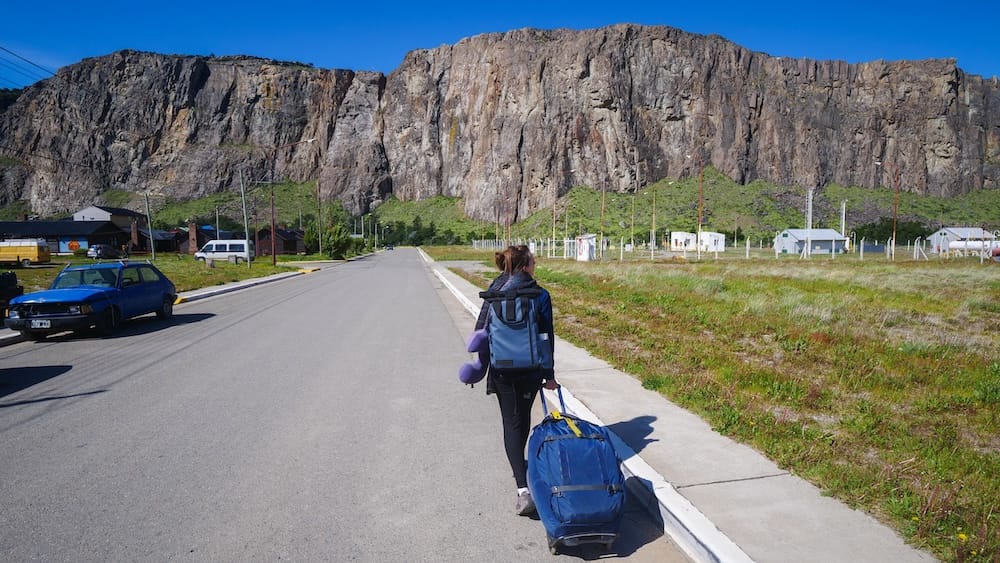 Arriving in El Chaltén, Patagonia, Argentina, with Luggage and Granite Cliffs, Featuring Audrey Bergner Audrey Bergner pulls a large wheeled duffel along a quiet street after arriving in El Chaltén, Santa Cruz, Patagonia, Argentina, with a backpack, roadside cabins and towering granite cliffs under a clear blue sky.