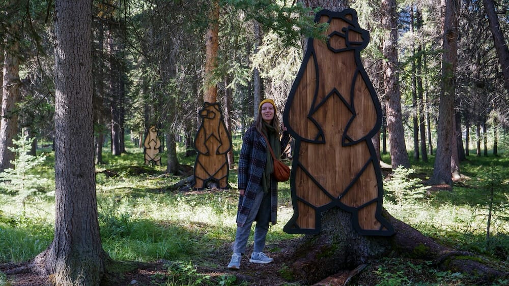 Audrey Bergner at Art in Nature in Banff town, Alberta, Canada beside wooden bear installations in the forest Audrey Bergner stands beside large wooden bear-shaped art installations along the Art in Nature trail in Banff town, Alberta, Canada, surrounded by conifer forest and filtered sunlight.