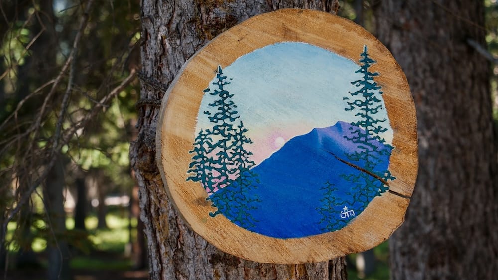 Outdoor art panel along the Bow River Trail, Banff, Alberta, Canada A round hand-painted wood panel depicting mountains, pine trees, and a sunset is mounted on a tree trunk along the Bow River Trail in Banff, Alberta, Canada, part of a seasonal outdoor art display.
