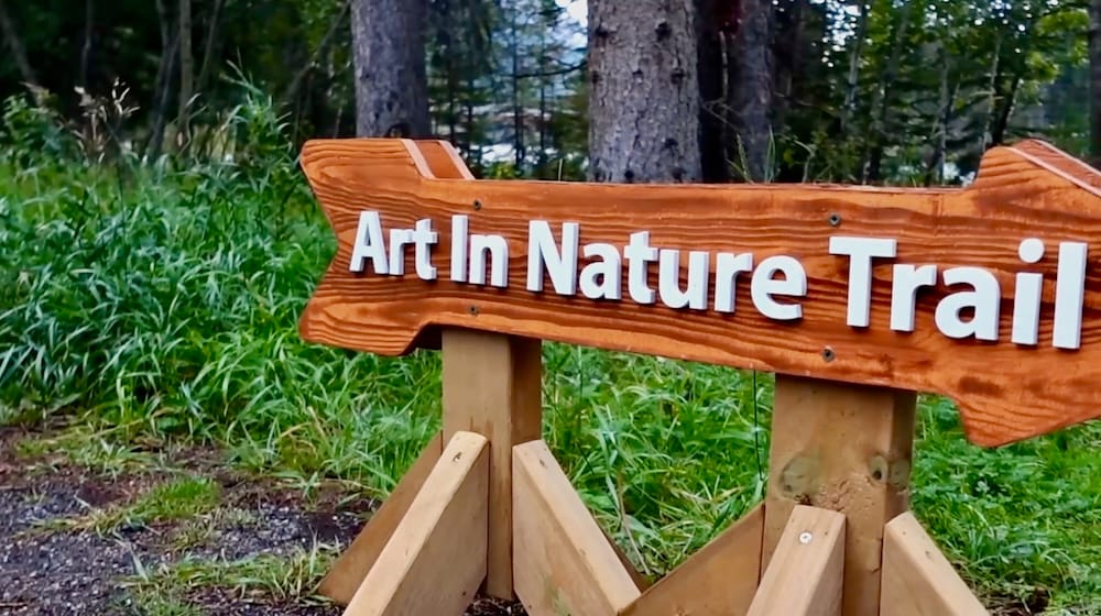 Art In Nature Trail trailhead sign, Banff, Alberta, Canada A wooden arrow-shaped sign reading "Art In Nature Trail" stands beside tall grass and trees at the start of the trail in Banff, Alberta, Canada.