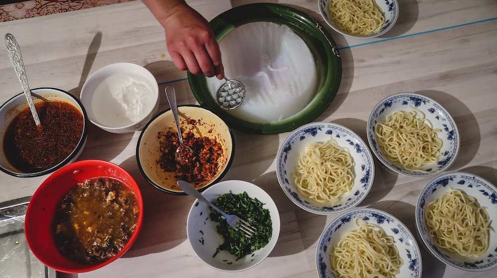 Bowls of wheat noodles are arranged beside translucent starch jelly, chilli sauce, chopped herbs and other condiments while a host prepares ashlan-fu during a Dungan family meal in Karakol, Kyrgyzstan.
