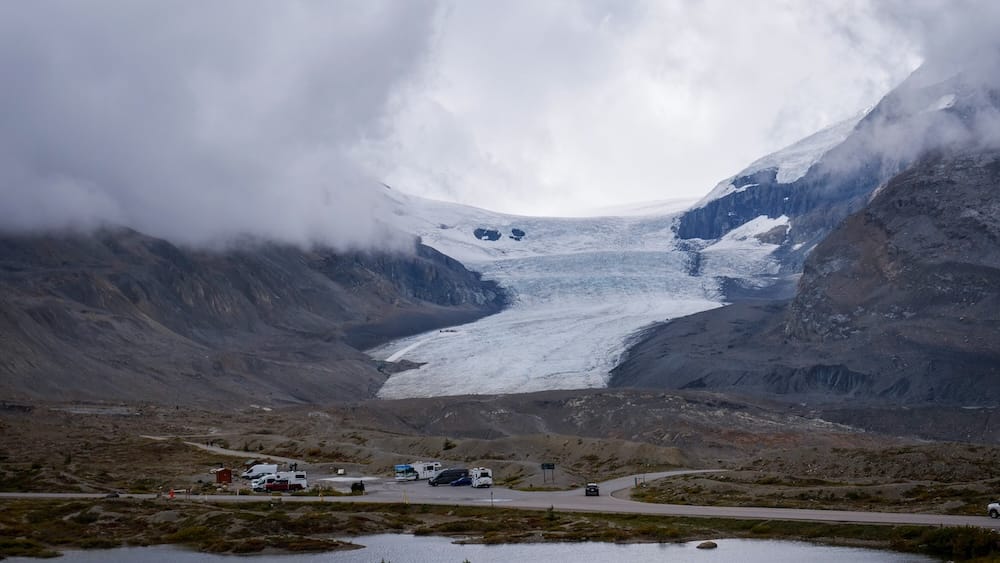 Campers, vans y otros vehículos estacionados cerca de la ruta bajo el glaciar Athabasca en Columbia Icefield, Alberta, Canadá, con el hielo descendiendo entre laderas rocosas bajo nubes densas.