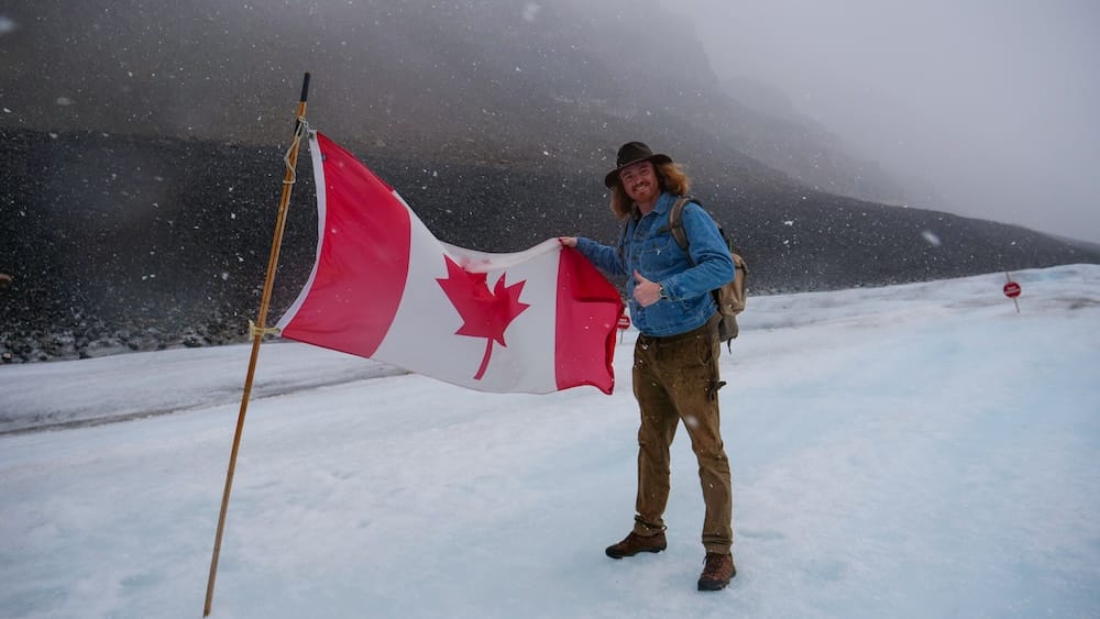 Samuel Jeffery está junto a una gran bandera canadiense sobre el glaciar Athabasca nevado en Columbia Icefield, Alberta, Canadá, mientras cae nieve en septiembre y la niebla oculta la morrena oscura y las laderas montañosas del fondo.
