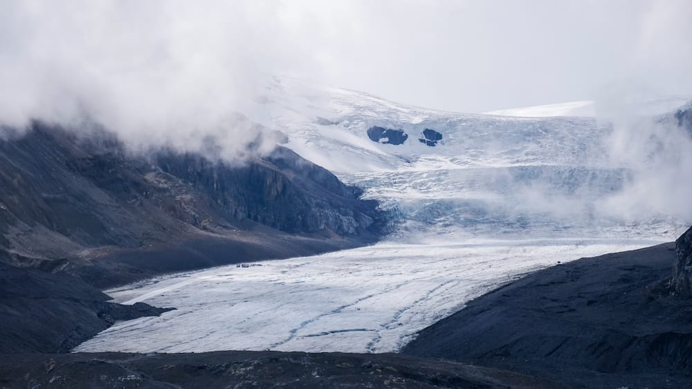 El glaciar Athabasca atraviesa morrenas oscuras y terreno montañoso en Columbia Icefield, Alberta, Canadá, con nubes bajas sobre el hielo superior que ocultan parcialmente las cumbres.