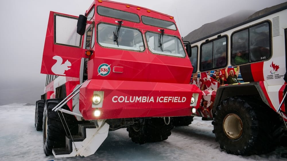 Un Ice Explorer rojo con la escalera extendida está sobre el glaciar Athabasca en Columbia Icefield, Alberta, Canadá, junto a otro vehículo glaciar, con hielo mojado y nubes bajas alrededor de la zona de ascenso.